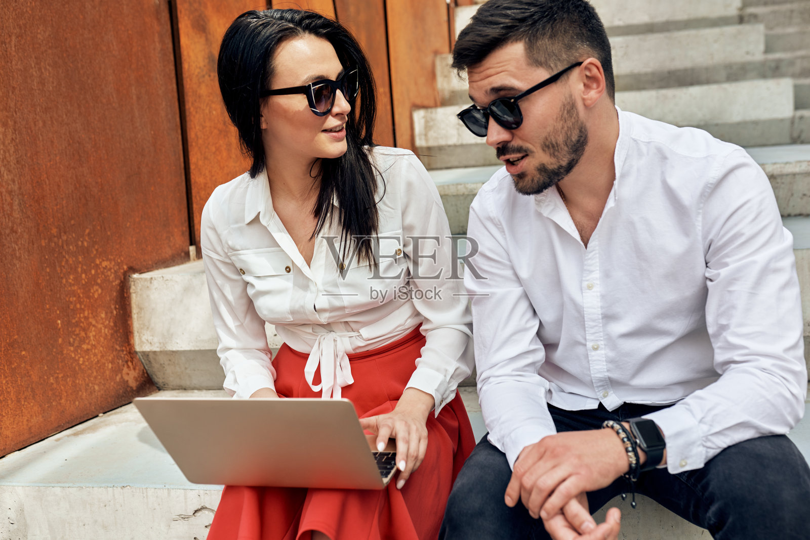 时尚的自由职业者使用科技进行户外工作。英俊的男人和微笑的女人坐在楼梯在一个城市中心穿着智能休闲装在笔记本电脑上一起工作。照片摄影图片