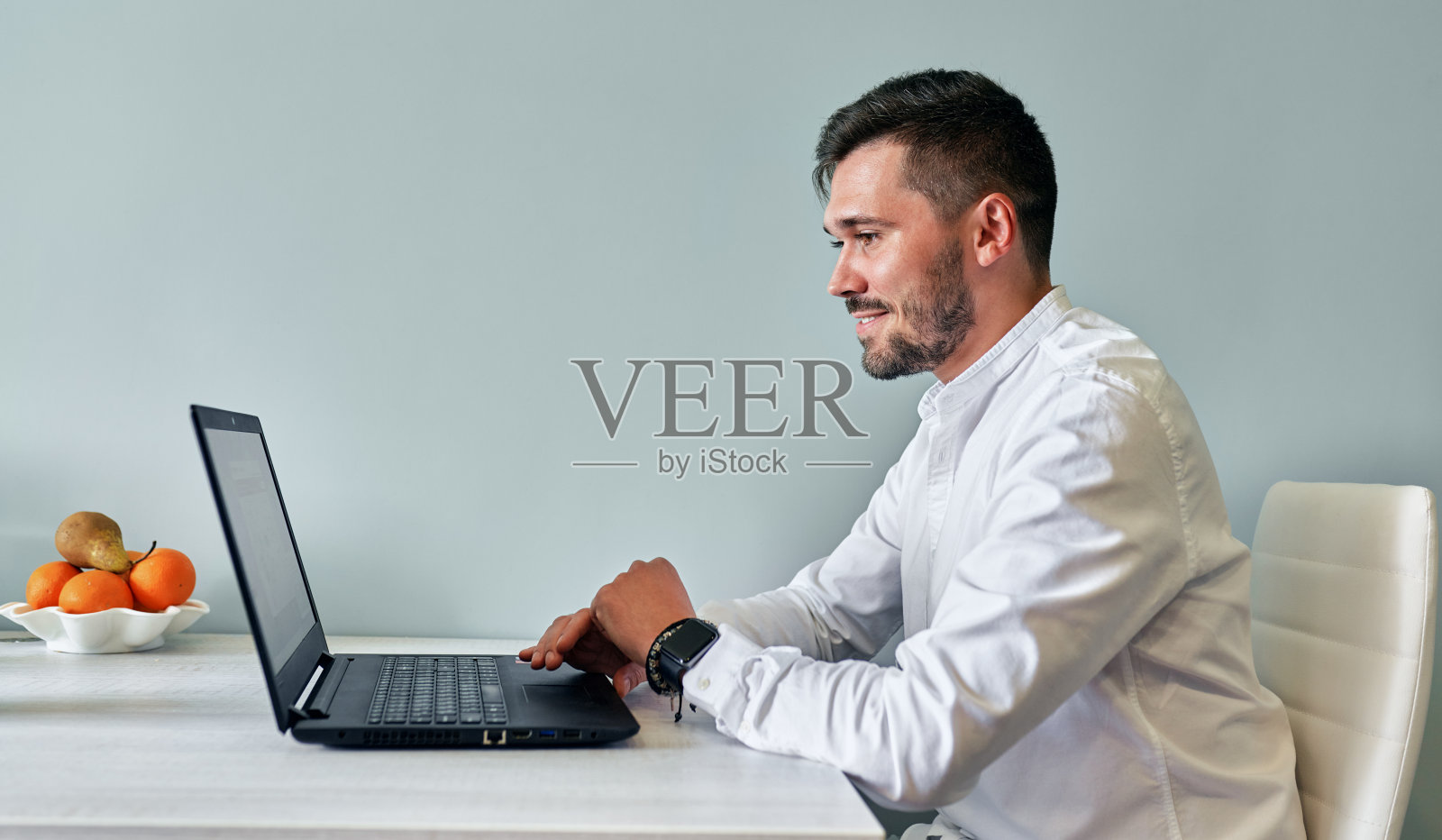 侧视图室内图像微笑的胡子男子工作在笔记本电脑上与朋友聊天，在线浏览。英俊的男性坐在厨房的桌子上，在电脑上工作。照片摄影图片