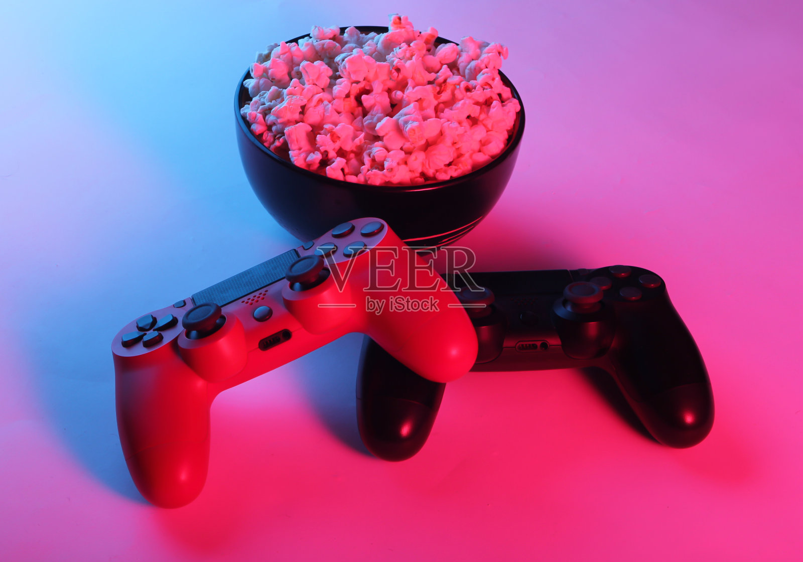 两个游戏手柄和一碗爆米花。霓虹粉蓝色的光。游戏、休闲和娱乐的概念。照片摄影图片