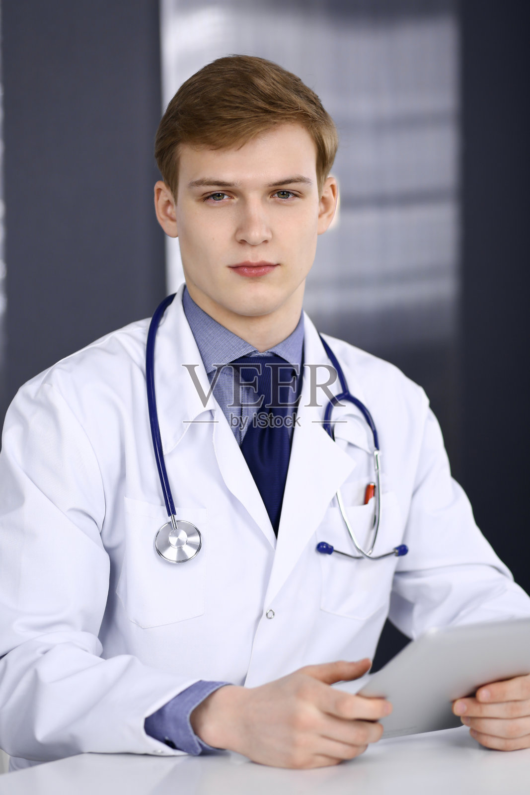 金发男医生坐在他工作地点的诊所里用平板电脑工作，特写。年轻医生在工作。完善的医疗服务，医学理念照片摄影图片