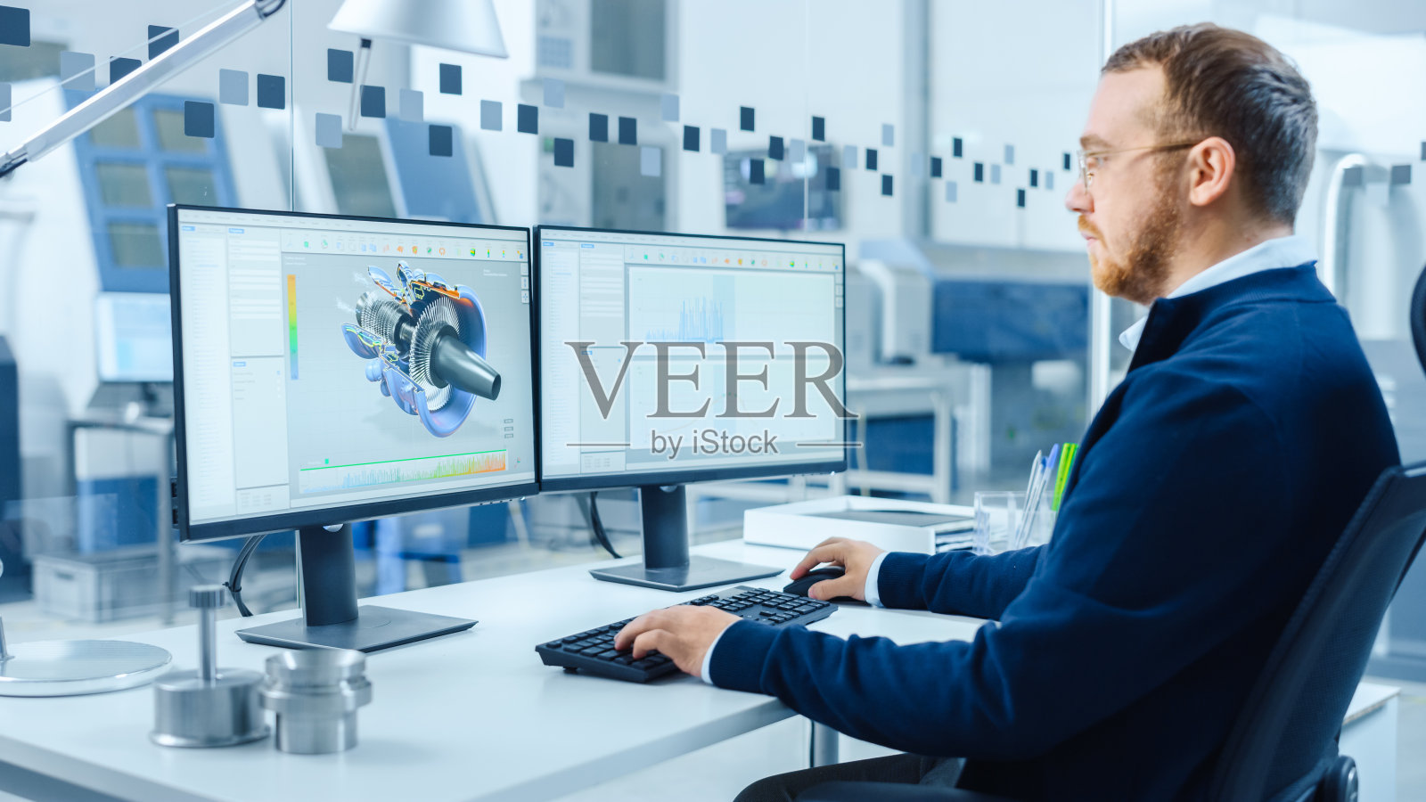 在个人电脑上工作的工业工程师，两个显示器屏幕显示CAD软件与3D原型的混合动力发动机和图表。拥有高科技机械的现代化工厂照片摄影图片