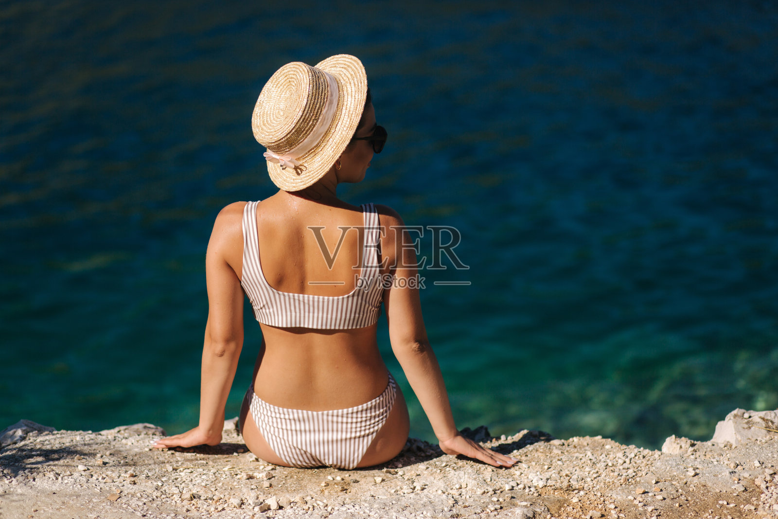 戴着帽子的漂亮年轻女子坐在美丽的泻湖边上的悬崖上。性感女人坐在海边的后视图。蓝色的水的背景。克罗地亚照片摄影图片