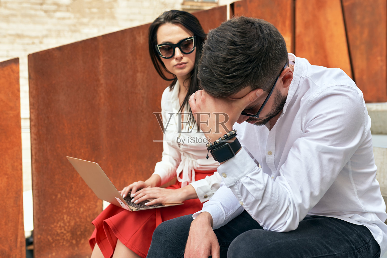 商业让那些使用科技在户外工作的人感到失望。一个悲伤的男人和严肃的女人坐在城市中心的楼梯上，穿着时髦的休闲服装，在笔记本电脑上一起工作照片摄影图片