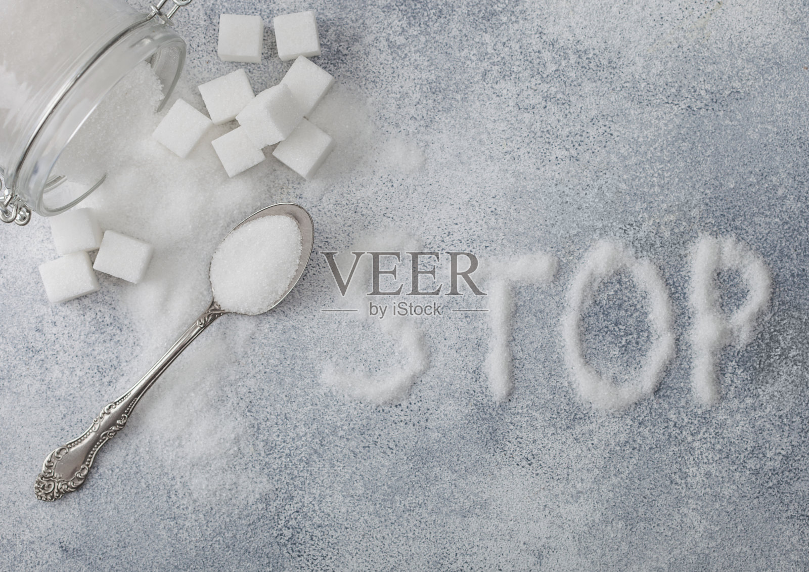 玻璃罐的天然白砂糖立方与银匙在光表背景与停止字母。不健康食品的概念。照片摄影图片