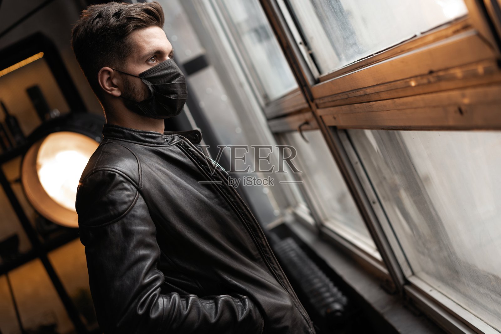 帅气的年轻人戴着黑色的面具。穿着皮夹克的时尚男士站在窗前看照片摄影图片