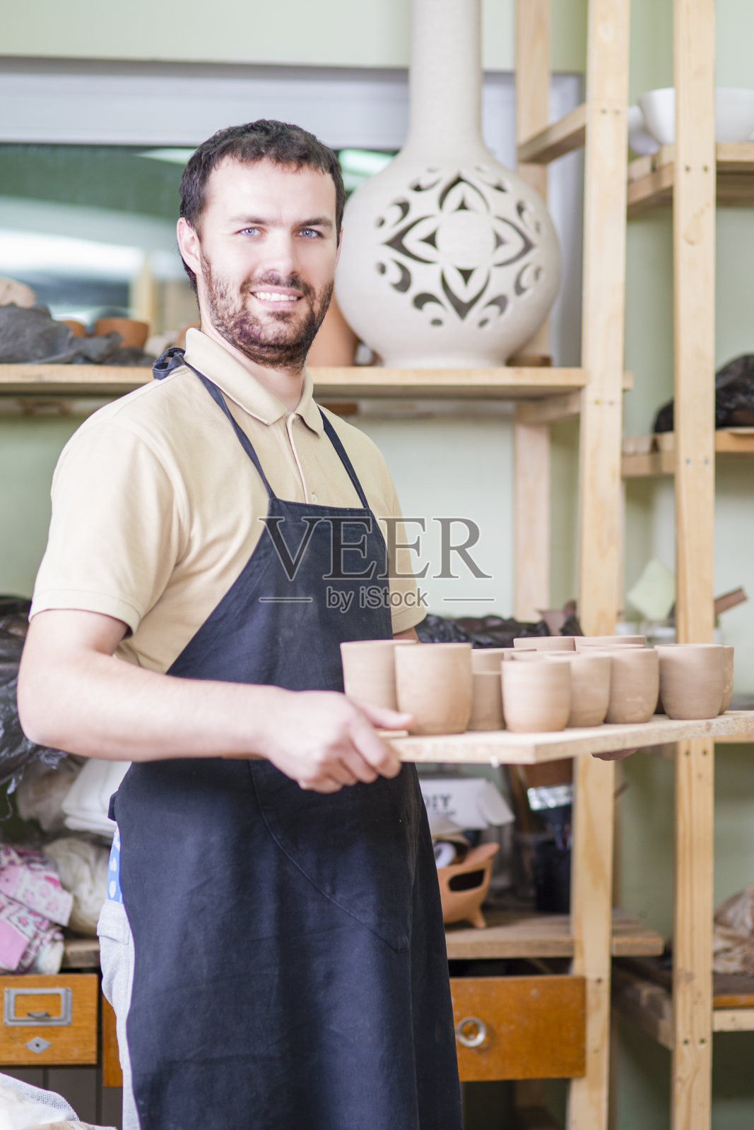专业男性陶艺家手持陶土手工杯子托盘的肖像。在车间的防护围裙中摆姿势。垂直的图像组成照片摄影图片