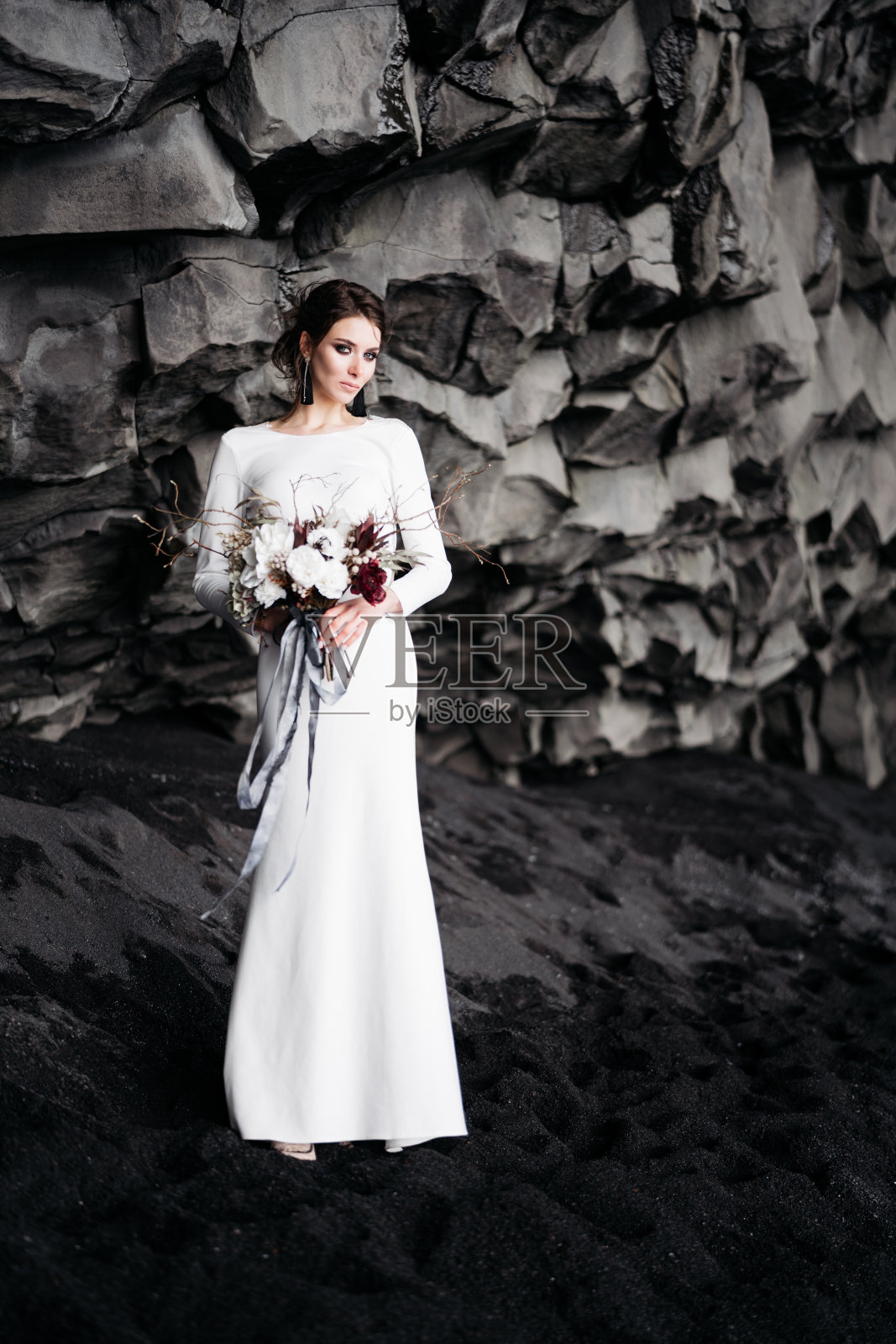 冰岛的目的地婚礼。新娘身穿白色丝绸礼服，手里拿着一束鲜花，站在维克海滩的黑色沙滩上，在玄武岩岩石山下。照片摄影图片