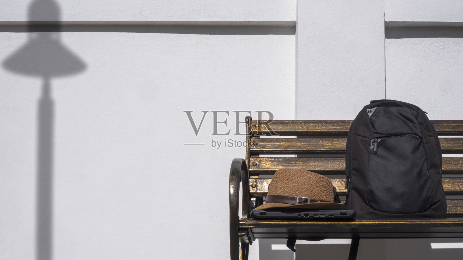 带笔记本电脑的软呢帽和背包放在公园的金属长椅上，公共区域的白色水泥墙背景上有路灯的影子照片摄影图片