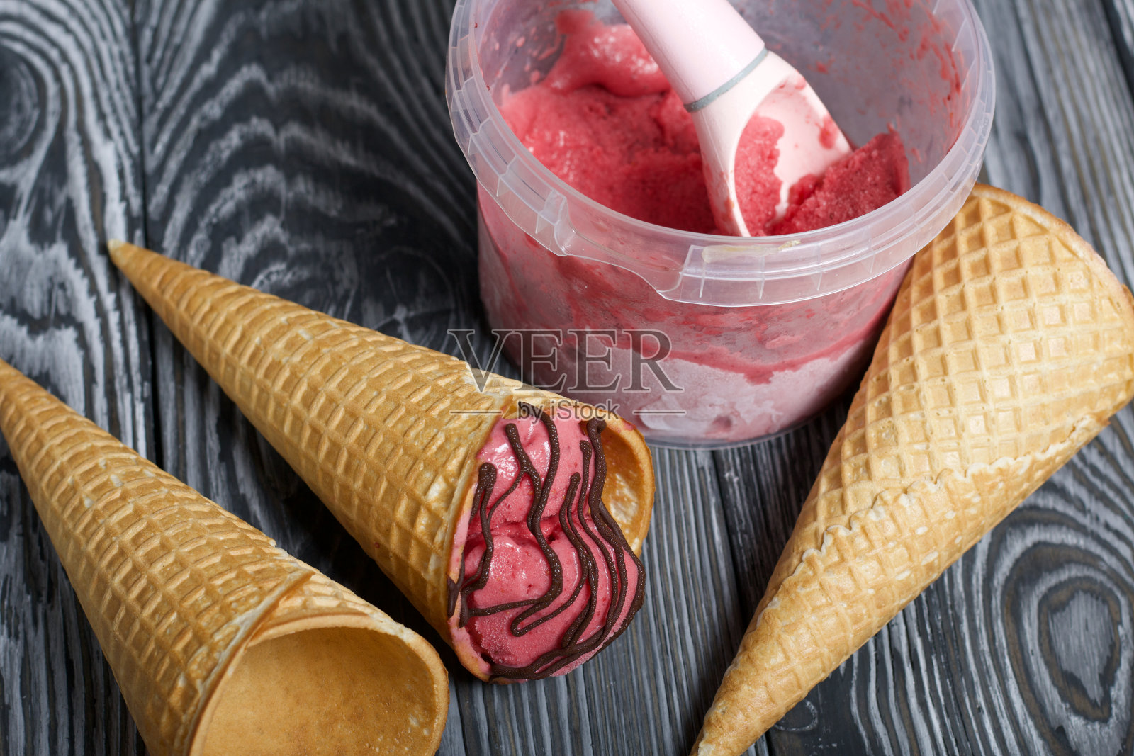 华夫蛋筒装草莓冰淇淋。配上巧克力。附近是空的没有冰淇淋的华夫蛋筒。冰淇淋容器和勺子。在黑色和白色的松木板上。照片摄影图片