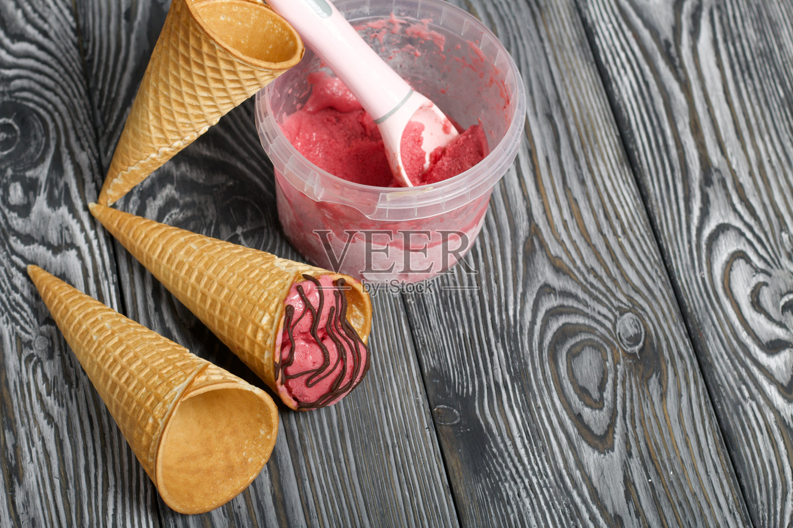 华夫蛋筒装草莓冰淇淋。配上巧克力。附近是空的没有冰淇淋的华夫蛋筒。冰淇淋容器和勺子。在黑色和白色的松木板上。照片摄影图片