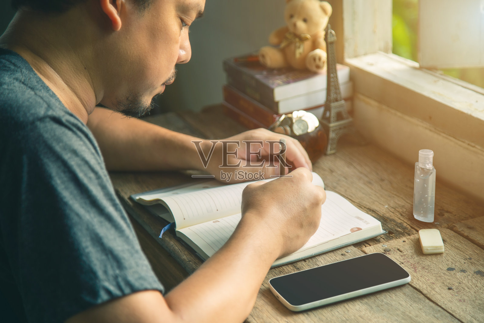 近距离观察一名亚洲男子，他穿着灰色t恤，坐在窗户旁边的一张旧木桌上，用手机写日记。照片摄影图片