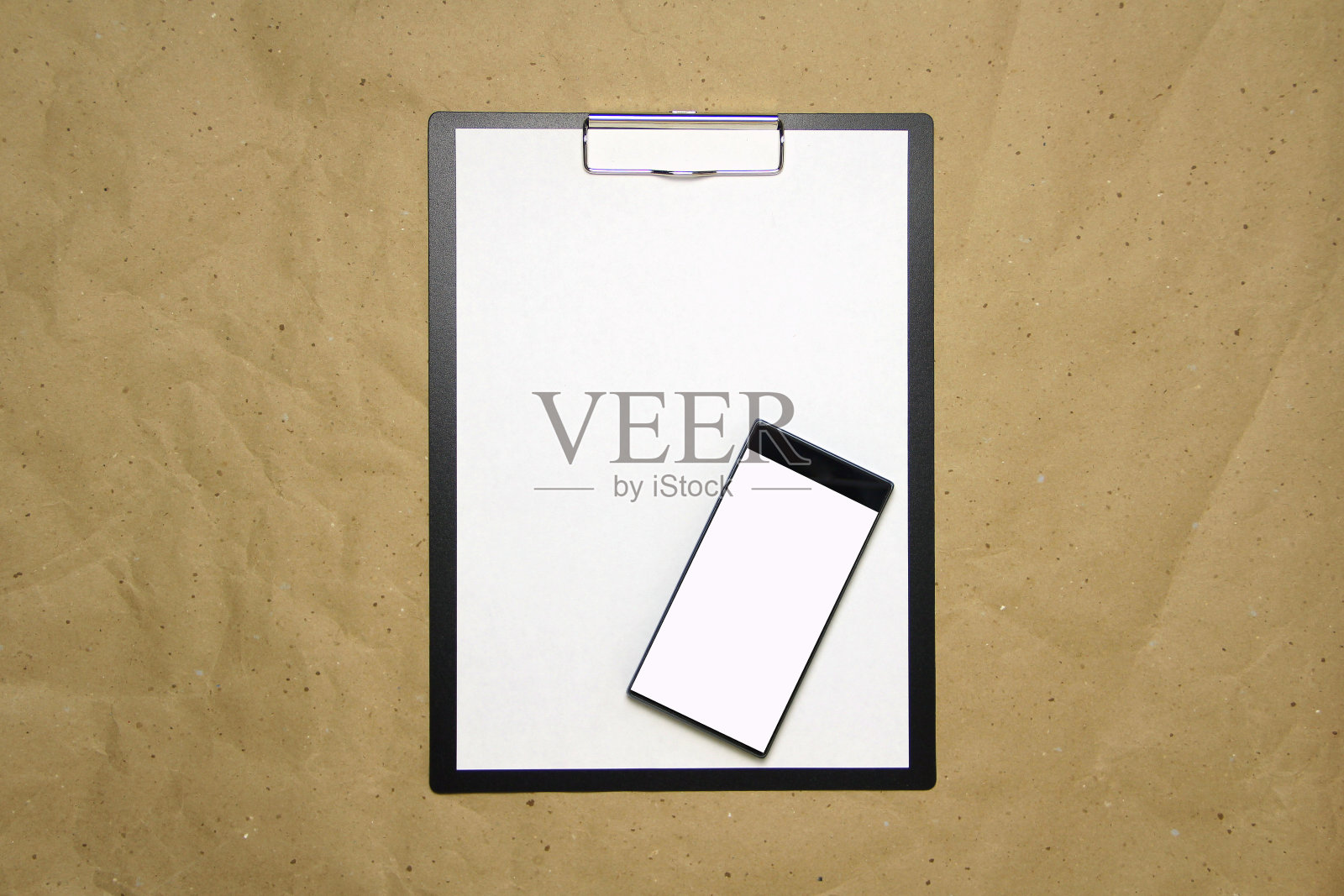 tablet with a clip for The clip of paper with a white sheet a4 lies against The background of craft brown crumpled paper. tablet with a clip for The clip of paper with a white sheet a4的空白纸。上图是一台白色屏幕的手机。放置文本，复制空间和布局设计照片摄影图片