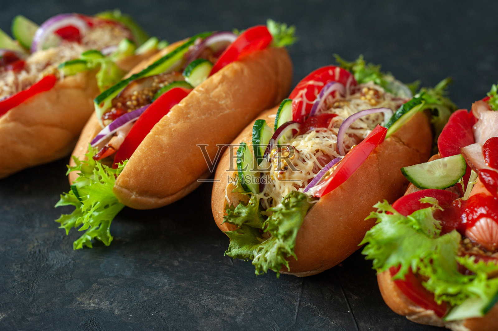 深色背景上的四个热狗。新鲜西红柿，黄瓜，生菜，香肠，培根，奶酪，芥末和番茄酱。照片摄影图片