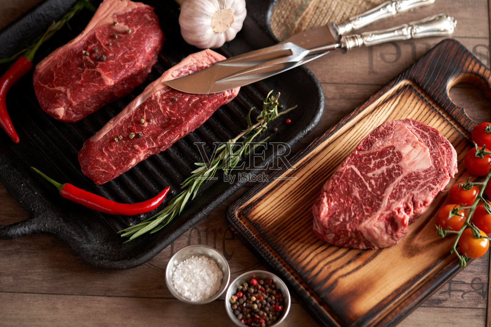 烤盘上的两块生肋眼牛排和砧板上的生肋眼牛排，加盐，花椒，红辣椒，大蒜头，迷迭香，一把肉叉和一把刀。烧烤肉照片摄影图片