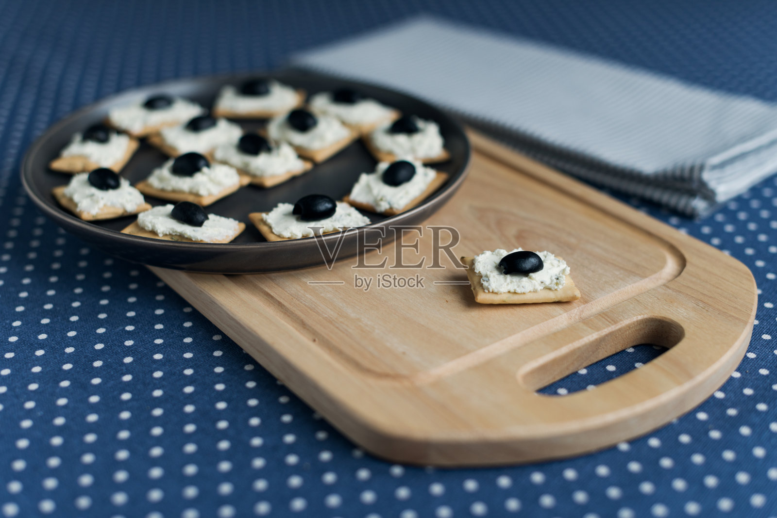 盘子上放有软奶酪和橄榄的饼干，铺在蓝色圆点桌布上。照片摄影图片