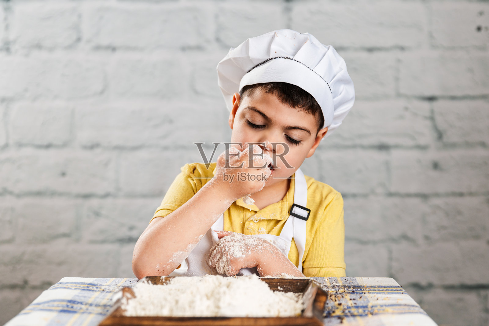 一个打扮成面包师的男孩把面粉送到嘴边，他用脏兮兮的手从面包、面粉和松饼堆里拿了起来。照片摄影图片