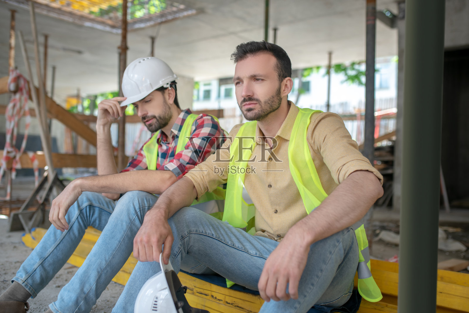 疲惫的建筑工人穿着黄色背心坐在木板上，其中一个手里拿着头盔照片摄影图片