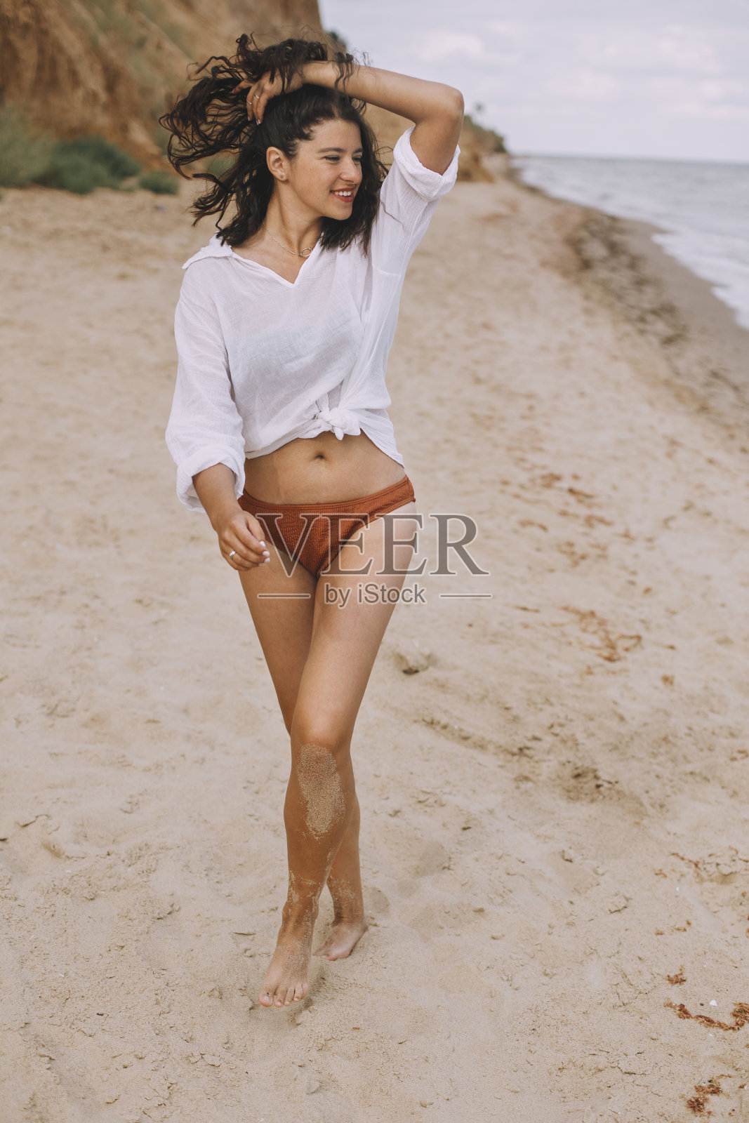 快乐的波西米亚女孩穿着白衬衫走在阳光明媚的海滩上。悠闲时尚的女人在泳装和衬衫放松在海边。暑假。生活真实的形象照片摄影图片