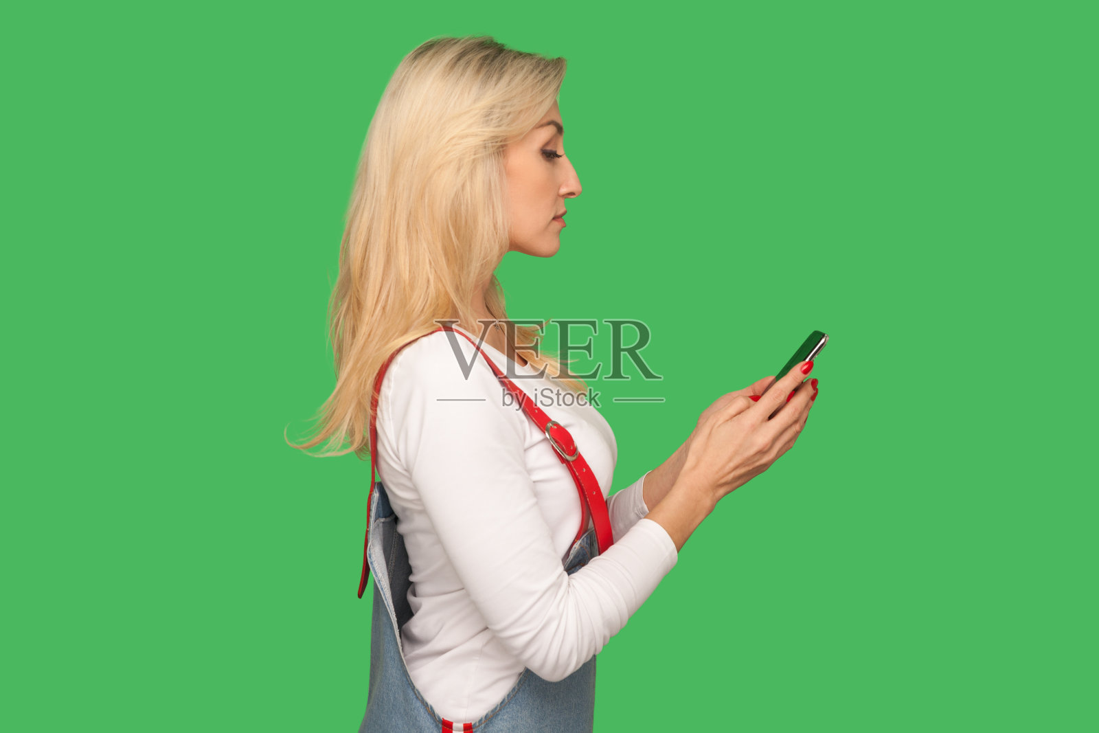 侧面的成年漂亮女人在时尚的牛仔工装裤拿着智能手机和阅读信息照片摄影图片