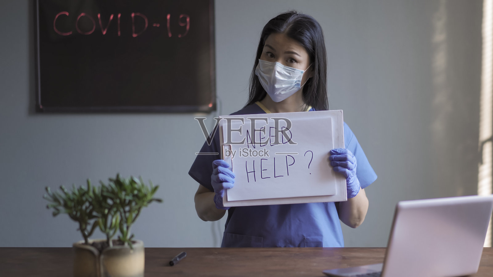女医生疑惑是否需要帮助，疑惑地看着摄像机。身穿医疗制服的亚洲妇女站在办公室里，背景是写有covid-19字样的黑板书照片摄影图片