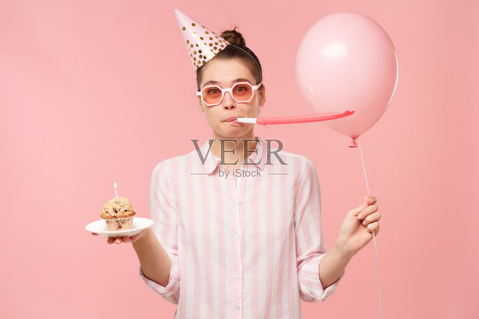 假日女孩戴着彩色眼镜和生日帽，拿着一个气球和盘子上的纸杯蛋糕，吹着口哨，孤立在粉红色的背景照片摄影图片