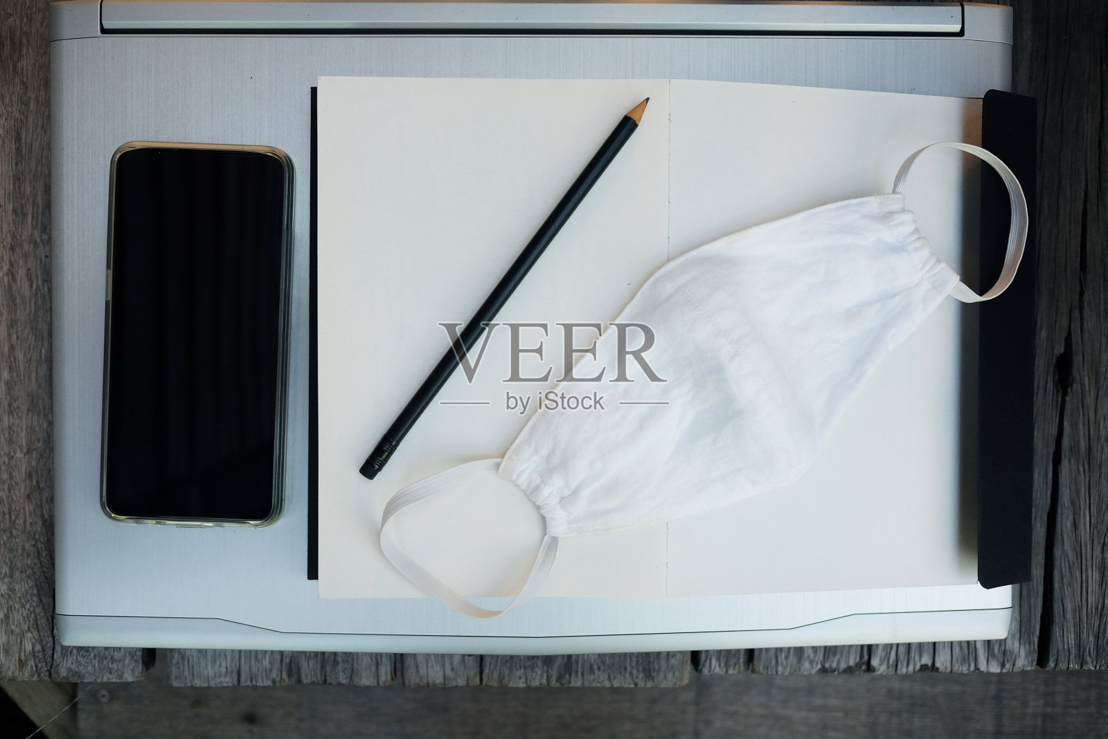 预防Covid-19或冠状病毒的白布口罩，用于商业技术的智能手机、书籍、笔记本电脑上的铅笔和在家办公的医疗理念。照片摄影图片