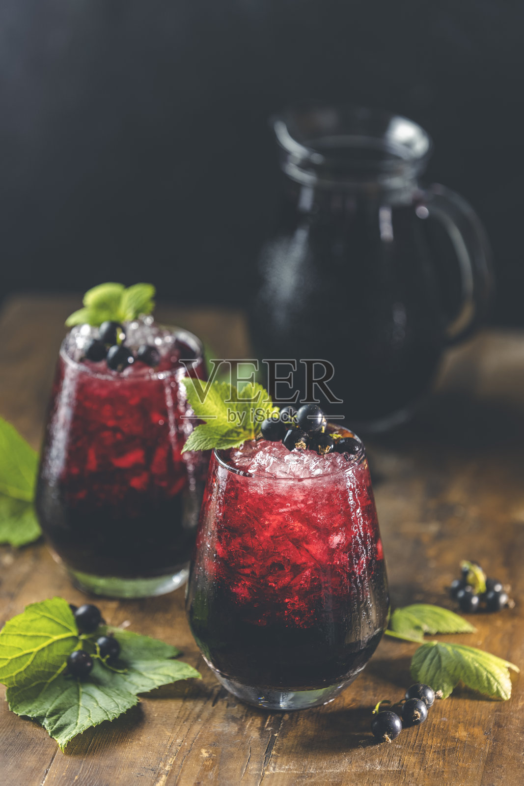 两杯冰黑加仑汁或鸡尾酒与成熟的浆果和绿叶在深色的木桌上。含酒精或不含酒精的夏季新鲜饮料照片摄影图片