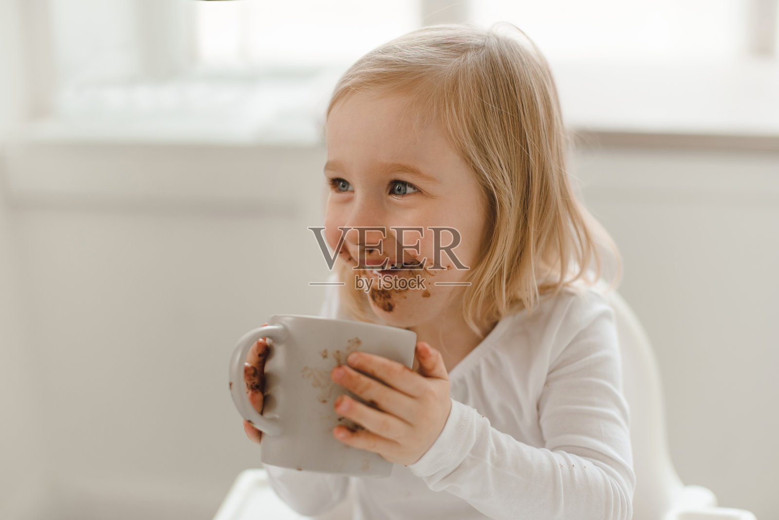 一个沾着巧克力的心满意足地微笑着的婴儿坐在桌子旁，手里拿着一个马克杯。照片摄影图片