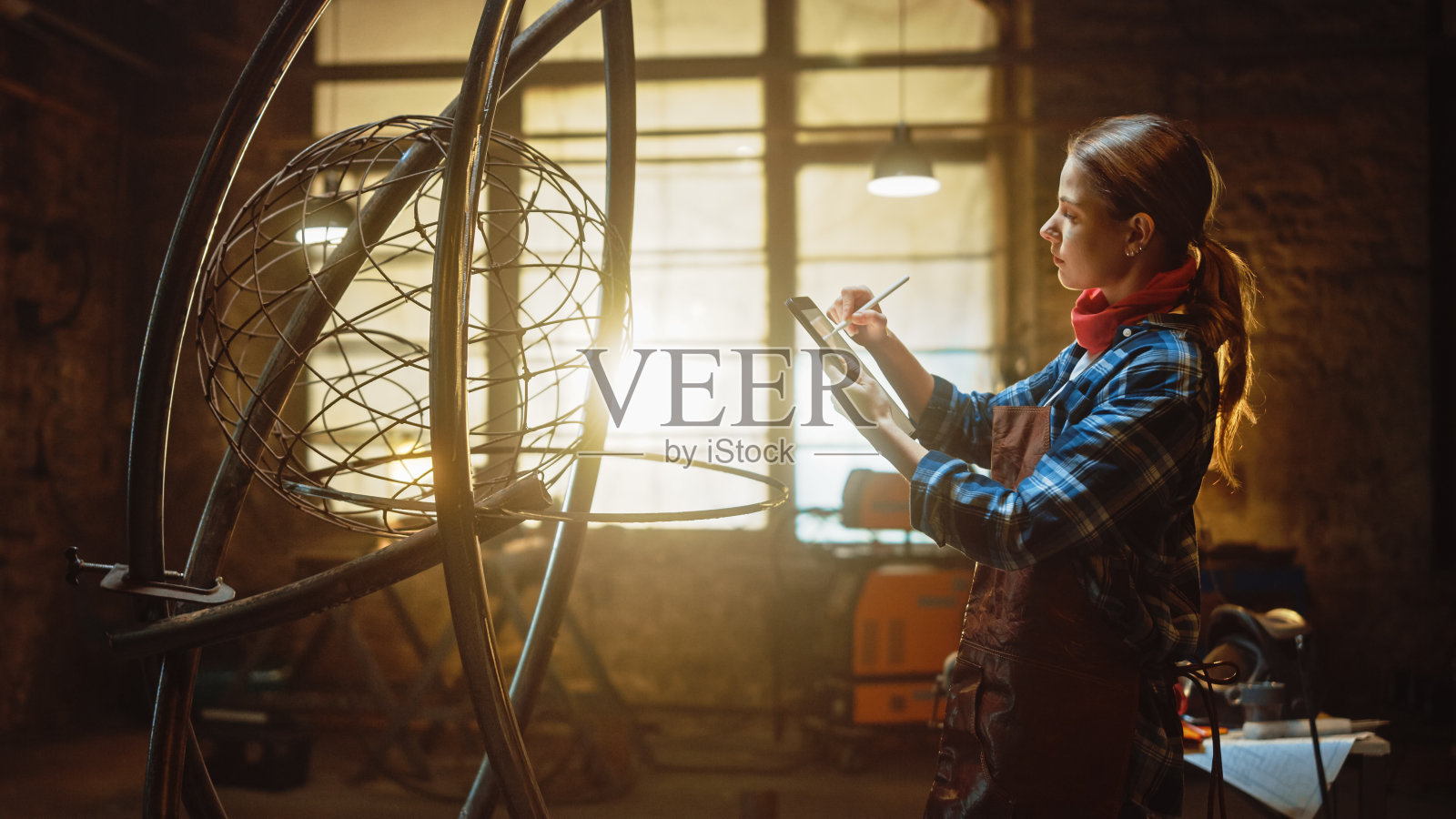 美丽的女性艺术家素描在平板电脑旁边的残酷的金属雕塑在工作室。假小子女孩穿着格子衬衫和围裙。当代制造者创造抽象的钢铁艺术。照片摄影图片