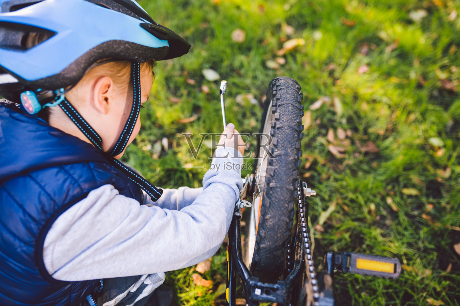 一个戴着头盔的白人男孩学会了修理他的自行车。骑单车的小孩在公园的草地上检查单车的机械装置。儿童运动俱乐部主题。山地车节照片摄影图片