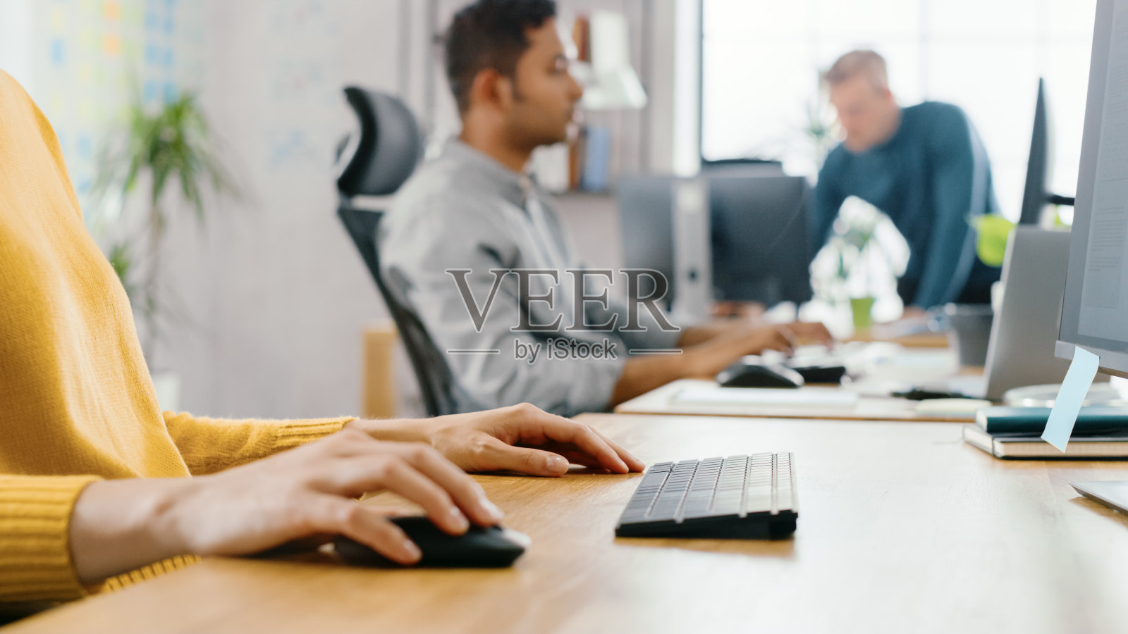 匿名创造性年轻女子的肖像坐在她的书桌上使用电脑鼠标和键盘。在背景明亮的办公室，多元化的年轻专业团队工作。照片摄影图片