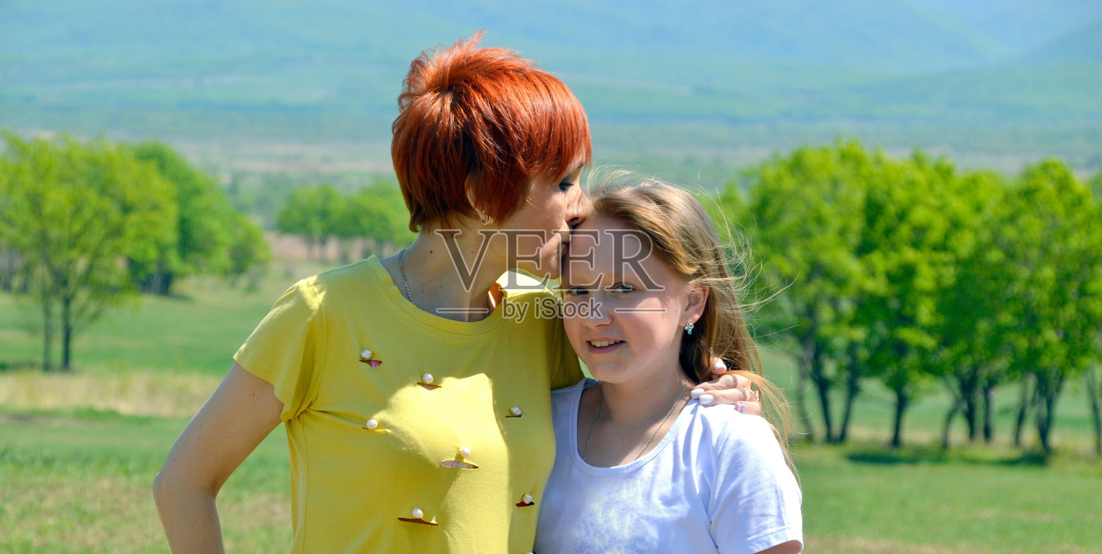 肖像的母亲亲吻她笑着女儿的前额在绿色的田野背景照片摄影图片