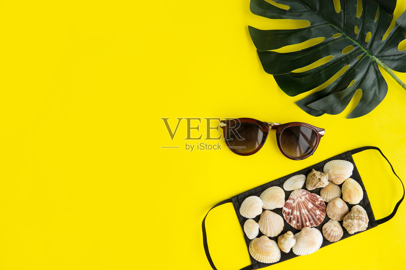 黑色医用口罩和贝壳，太阳镜，黄色背景上的怪物树叶。平躺、复印空间、度假取消、大流行、旅游。照片摄影图片