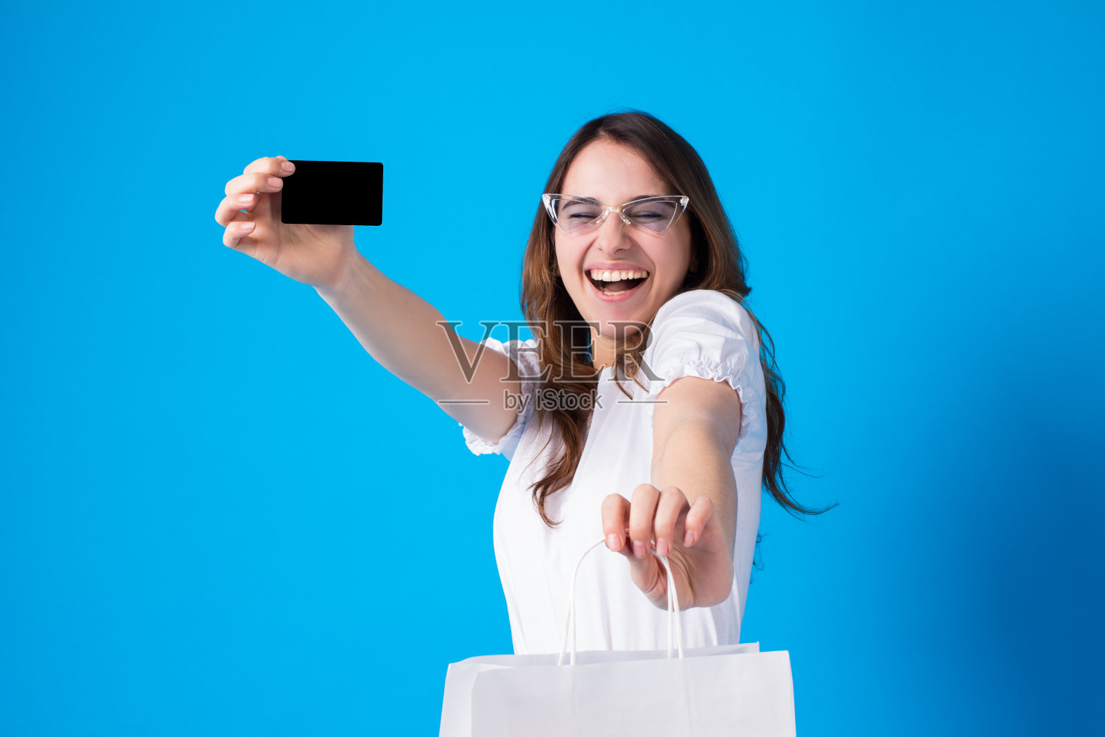 情绪激动微笑年轻漂亮的女人持有信用卡和白色工艺包装孤立在蓝色背景。穿着白色t恤和时尚太阳镜的女孩网上购物，银行，消费照片摄影图片