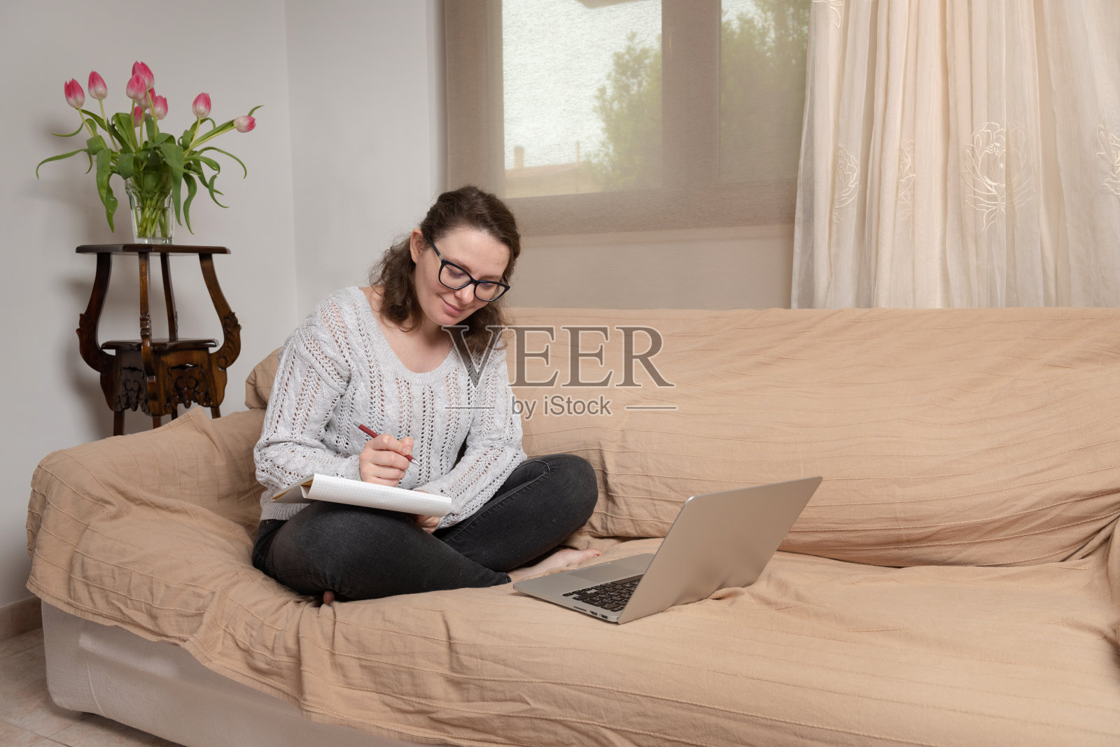 年轻女性坐在客厅的沙发上，在家远程工作。用她的笔记本电脑和书工作照片摄影图片