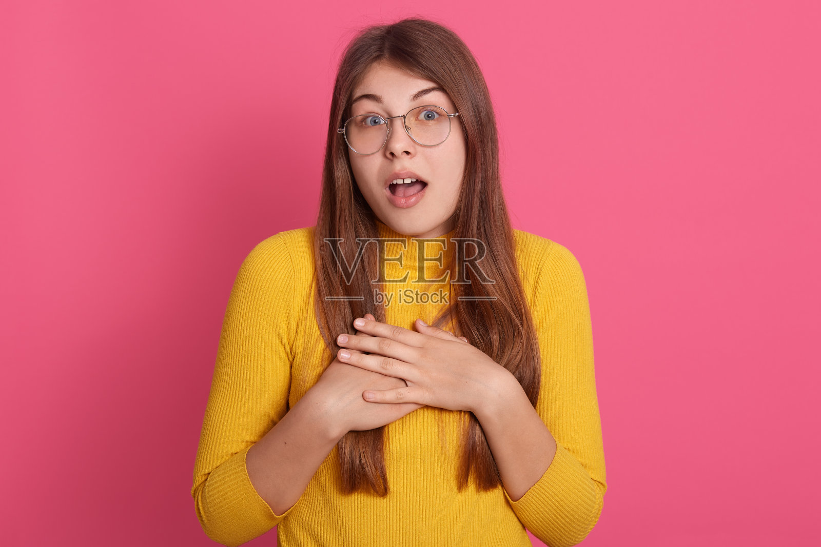 照片中，身着黄色运动衫，戴着眼镜的年轻女性情绪激动地直视着镜头，嘴巴和眼睛睁得大大的，面部表情震惊。情感的概念。照片摄影图片