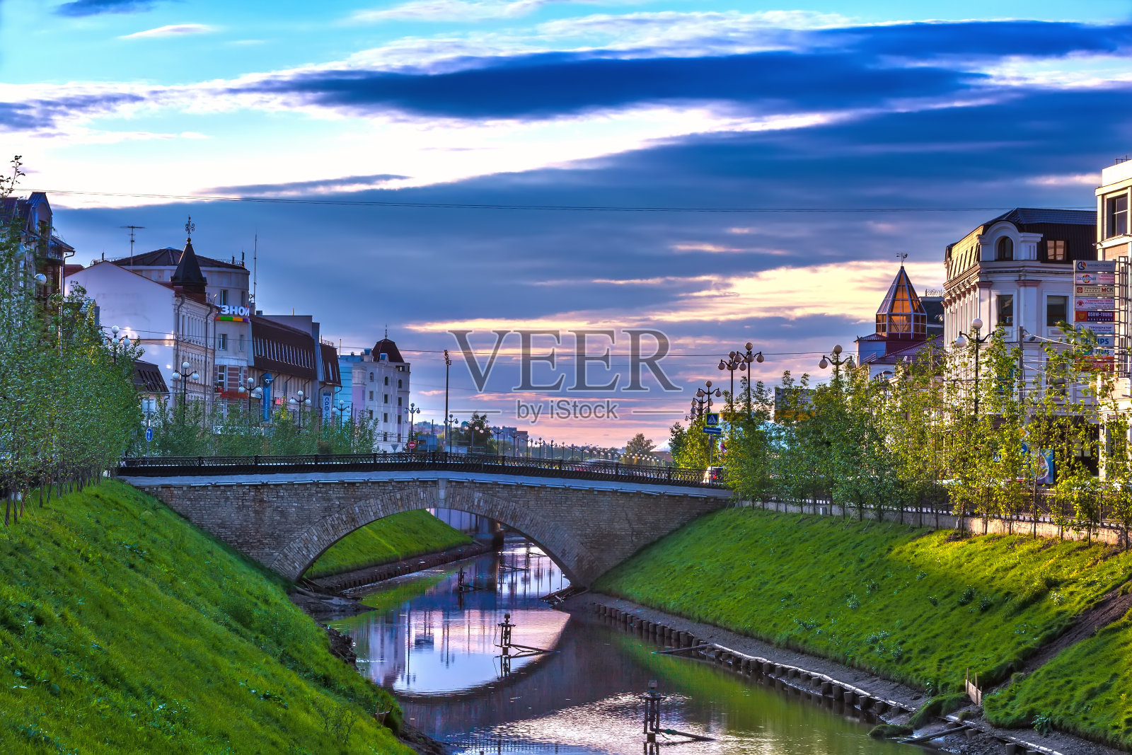 俄罗斯鞑靼斯坦喀山的布拉克管道。一条小小的城市河，在城市房屋之间，在夕阳的照耀下，有一个绿草如茵的斜坡照片摄影图片