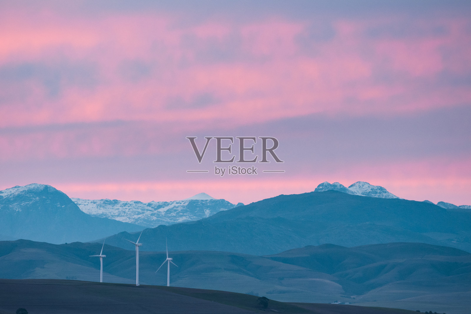 日落时分，白雪皑皑的山峰，前景中有风力涡轮机照片摄影图片