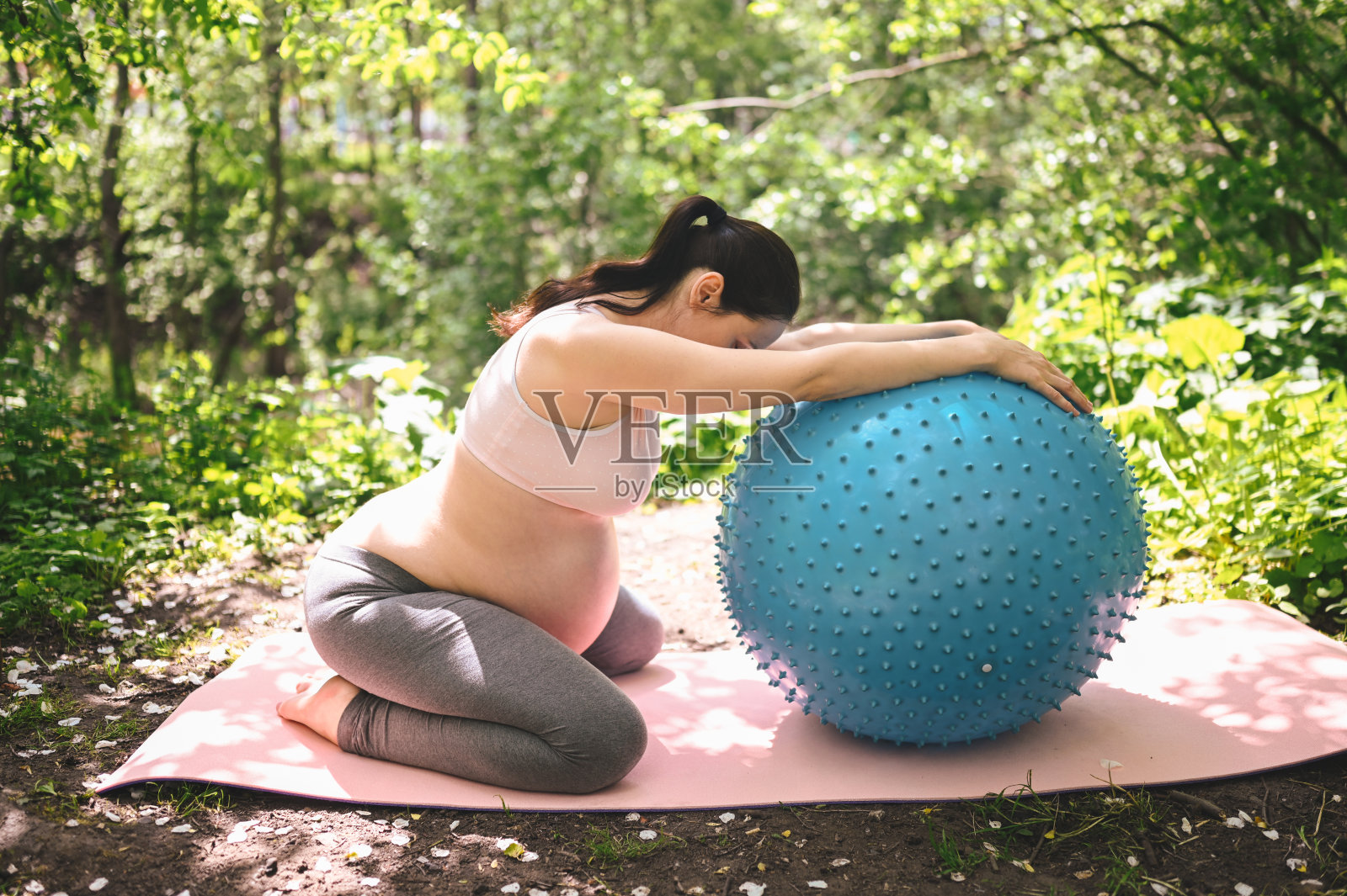 美丽的年轻孕妇做健身普拉提蓝球在公园户外。坐在粉红色的瑜伽垫上放松。积极的未来母亲运动生活方式。健康怀孕的概念照片摄影图片