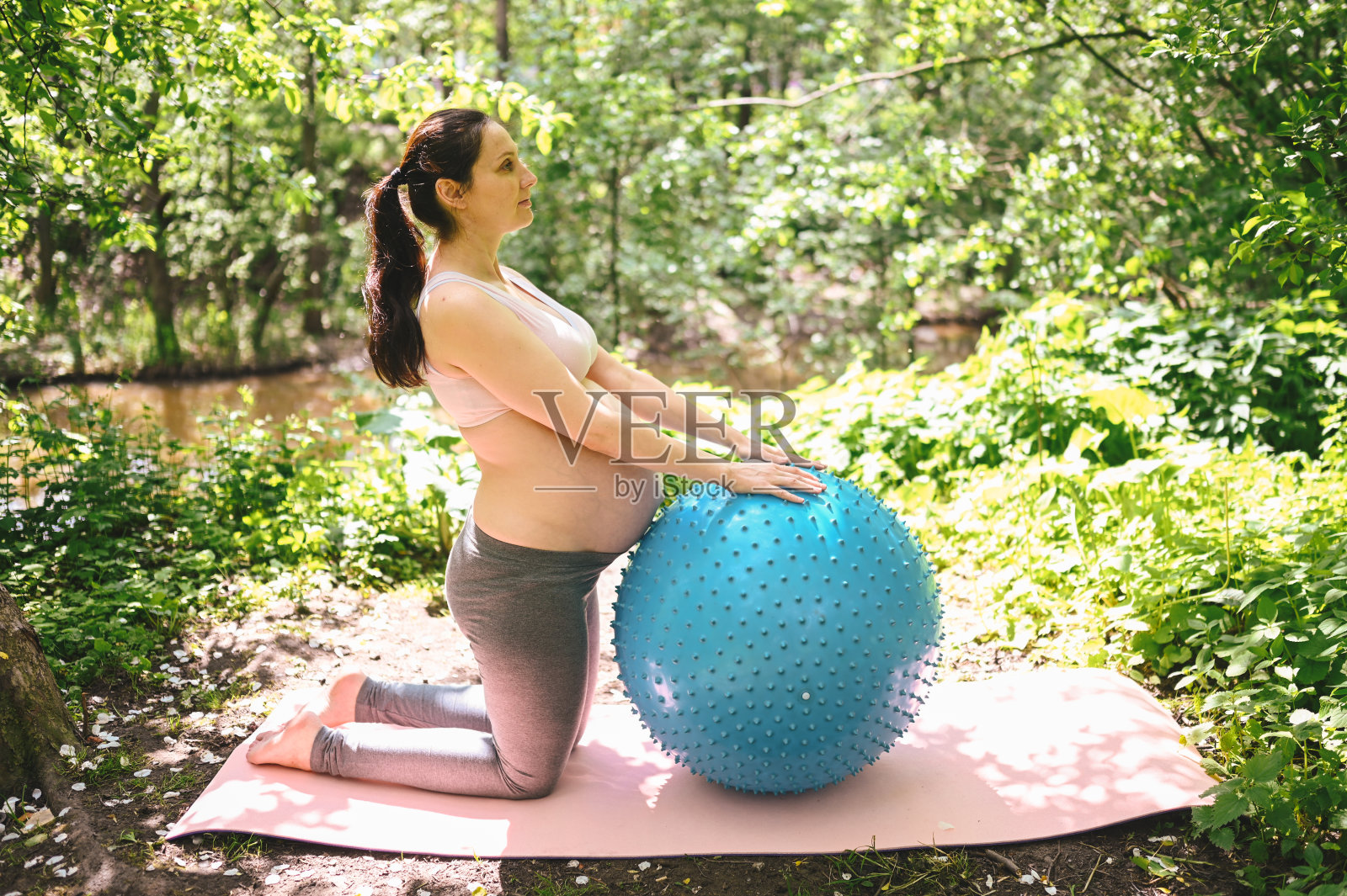 美丽的年轻孕妇做健身普拉提蓝球在公园户外。坐在粉红色的瑜伽垫上放松。积极的未来母亲运动生活方式。健康怀孕的概念照片摄影图片