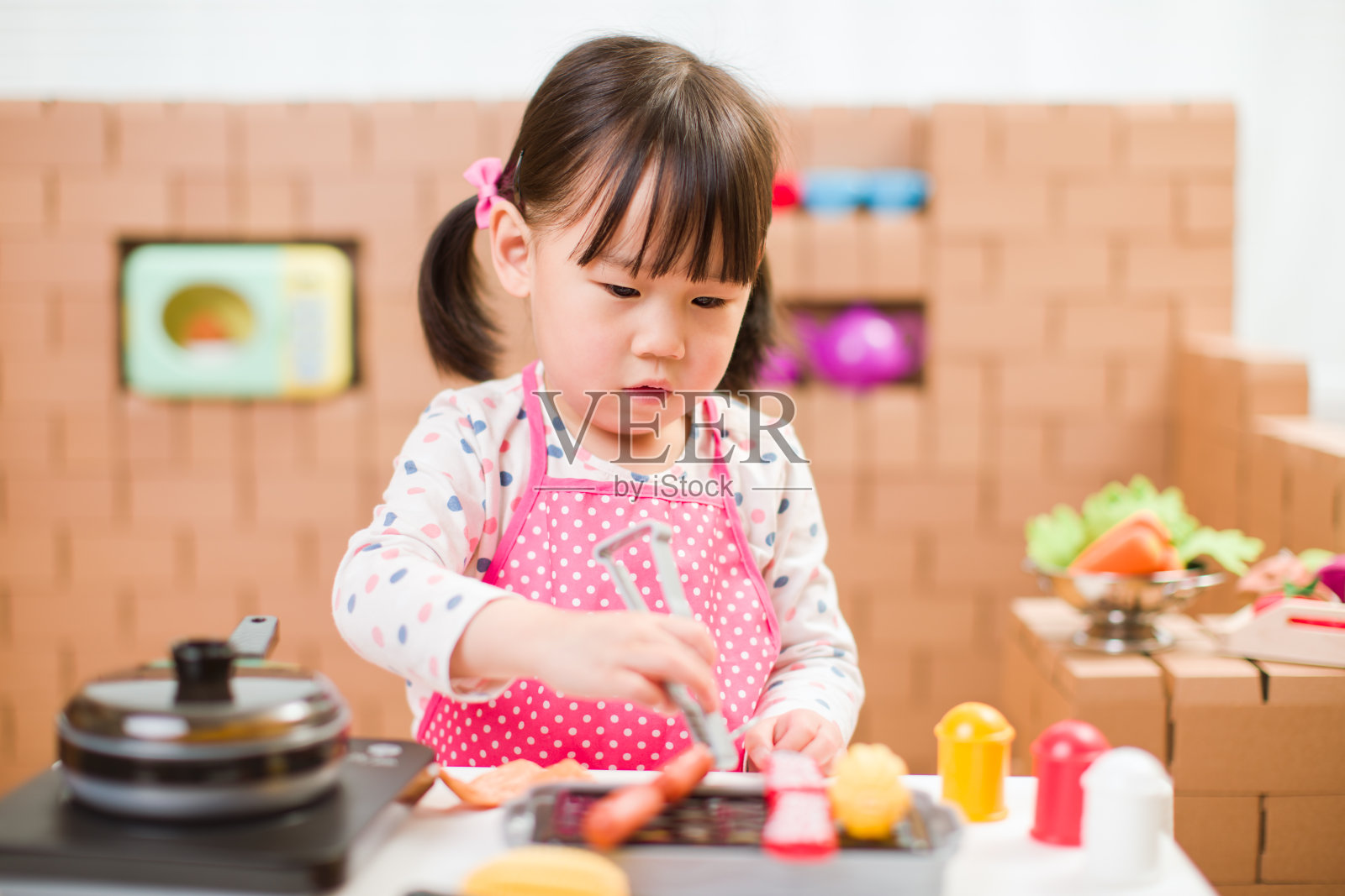 蹒跚学步的小女孩以纸板块为背景，假装扮演准备食物的角色照片摄影图片