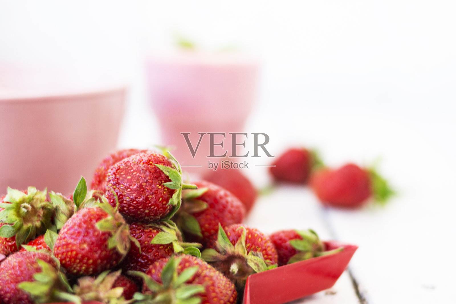 新鲜的草莓酸奶和浆果周围的白色木质背景。一个粉红色的奶昔在一个玻璃杯，旁边是一个粉红色的杯与赫拉克斯薄片，和红色的草莓躺在一个红色的星形盘子照片摄影图片