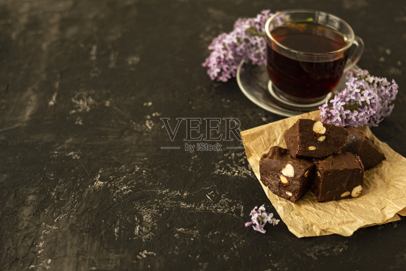 一块自制的巧克力蛋糕放在皱皱的牛皮纸上，黑色的桌子上放着坚果。一杯茶和丁香花装点着美味的茶会。照片摄影图片