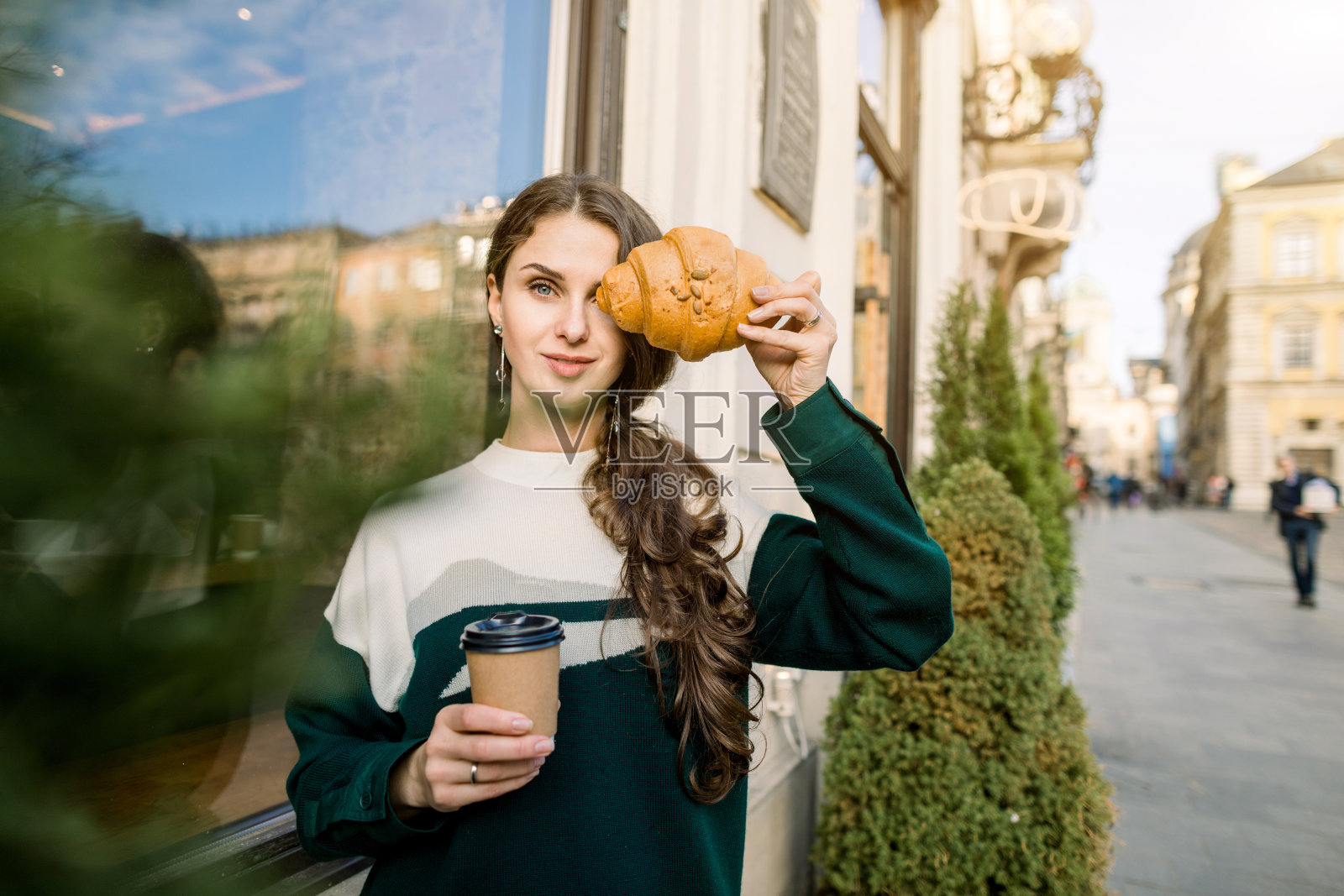 迷人的年轻女子对着镜头摆姿势，端着纸杯咖啡和新鲜美味的羊角面包，站在户外，背景是古老的城市建筑。照片摄影图片