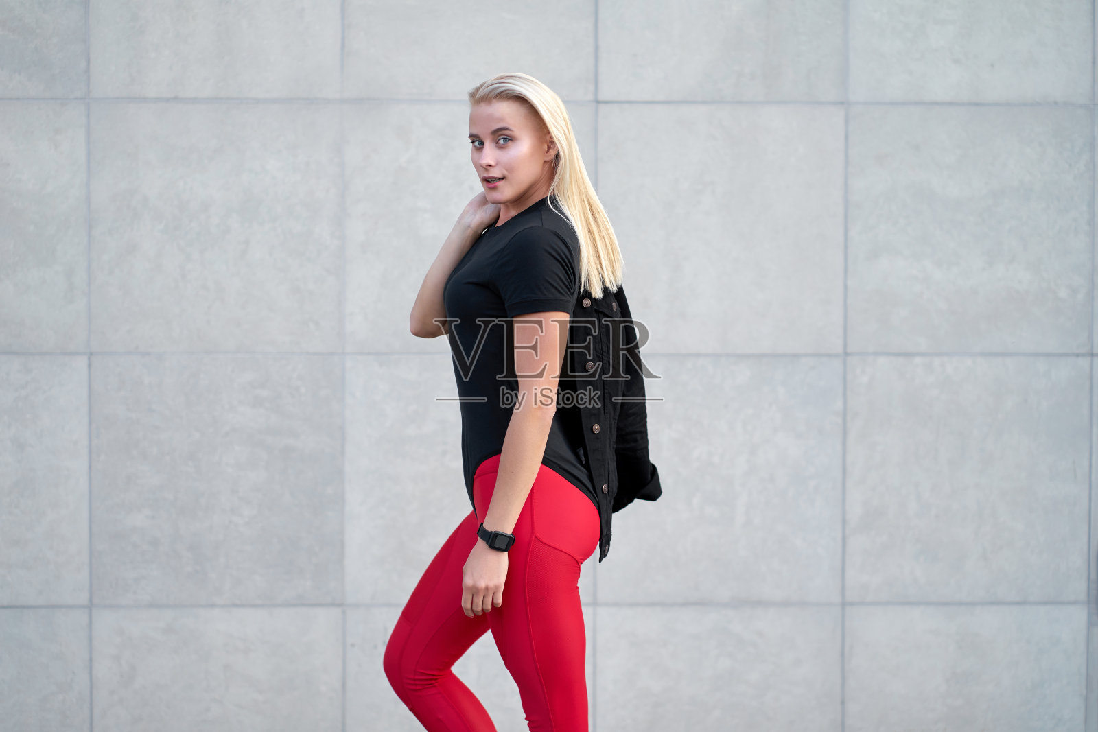 年轻迷人的运动女孩或模特的肖像穿着黑色运动服，时髦的红色打底裤。室外拍摄在灰色的墙壁背景。照片摄影图片