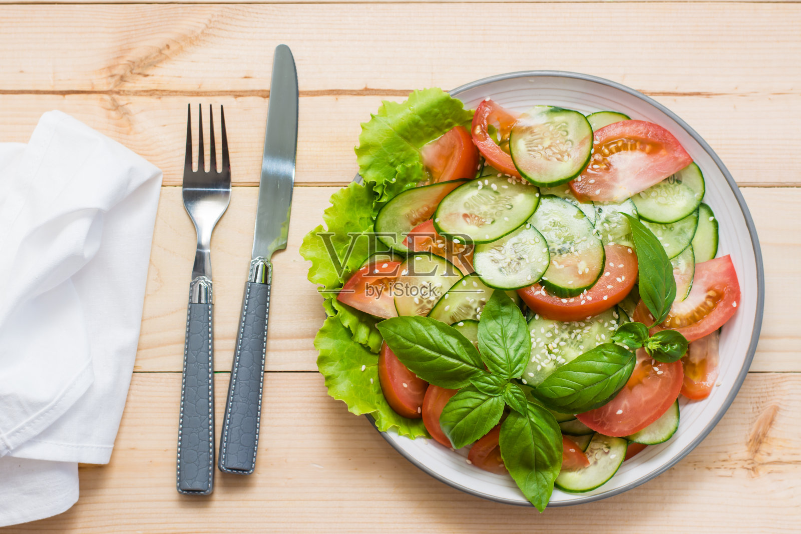传统的沙拉是把黄瓜片、西红柿和芝麻放在盘子里，用刀叉放在木桌上。素食。俯视图照片摄影图片