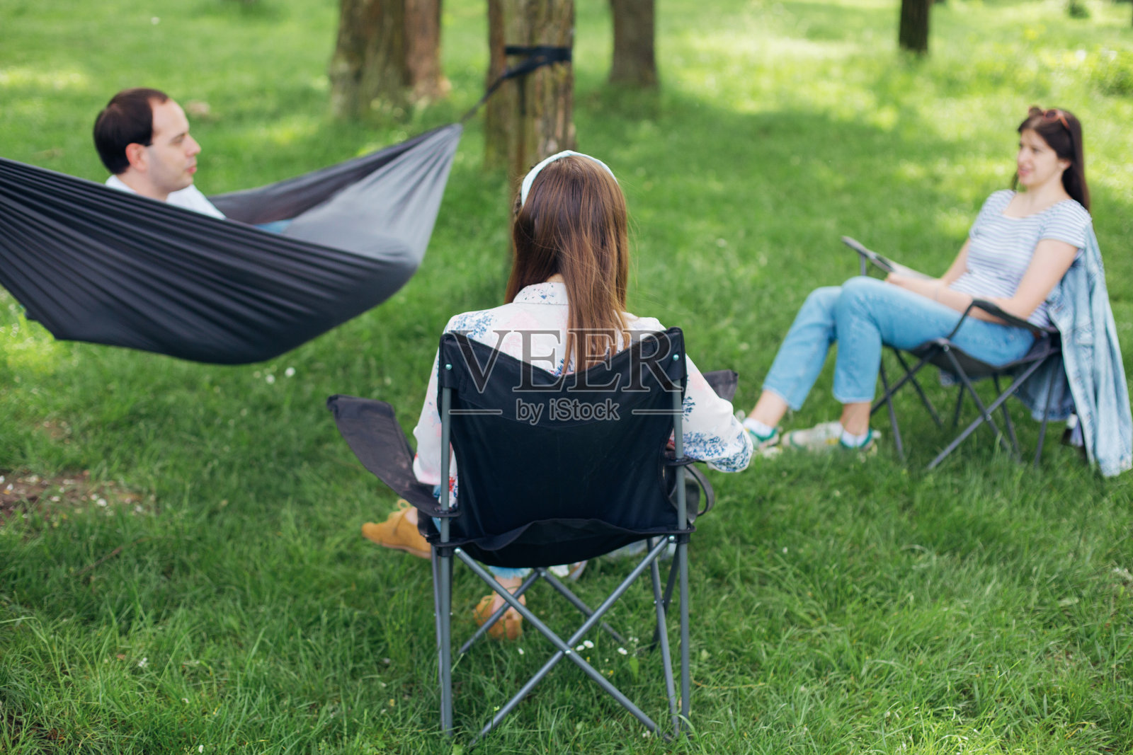社会距离。一小群人享受谈话在野餐与社会距离在夏季公园。朋友们在树间的吊床和椅子上休息。新常态，安全集会照片摄影图片