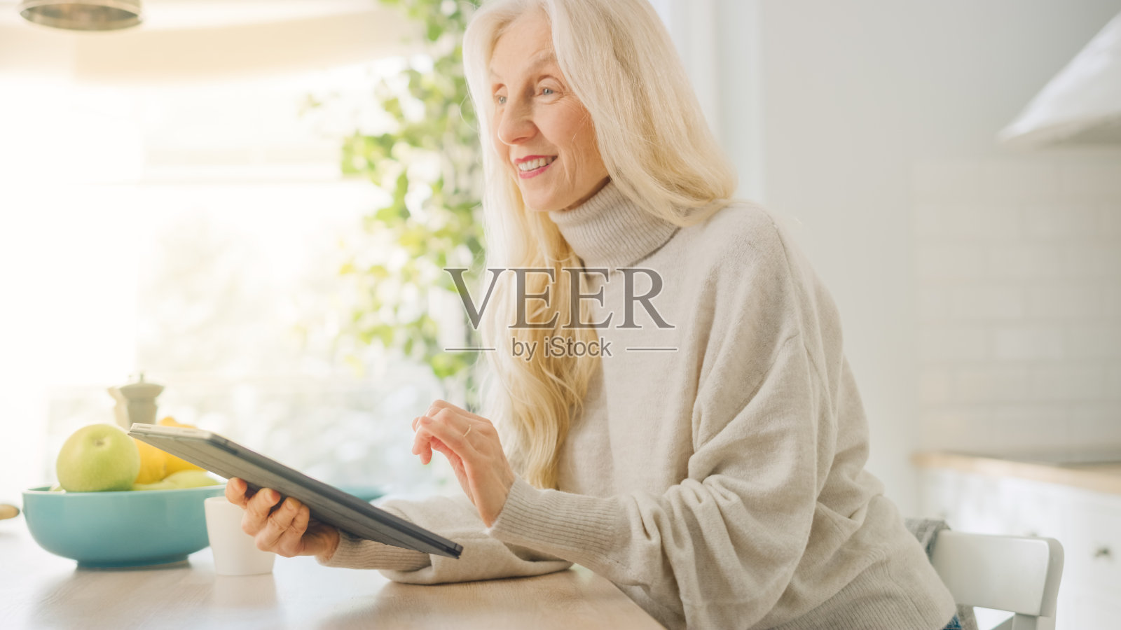 真正的高级女性在厨房用平板电脑在家。美丽的老年女性退休者与灰色头发写在一个设备上的信使文本。与科技携手的老年人。照片摄影图片