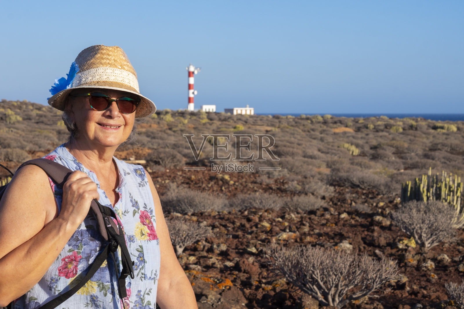 成年老妇人微笑着欣赏以灯塔为背景的海景。帽子和背包。干旱的热带地区，有火山岩照片摄影图片