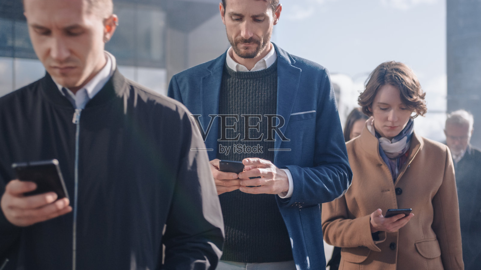 办公室经理和商人正在一座现代玻璃办公大楼前使用他们的智能手机。人们穿着得体，看起来很成功。他们被自己的设备占用。照片摄影图片